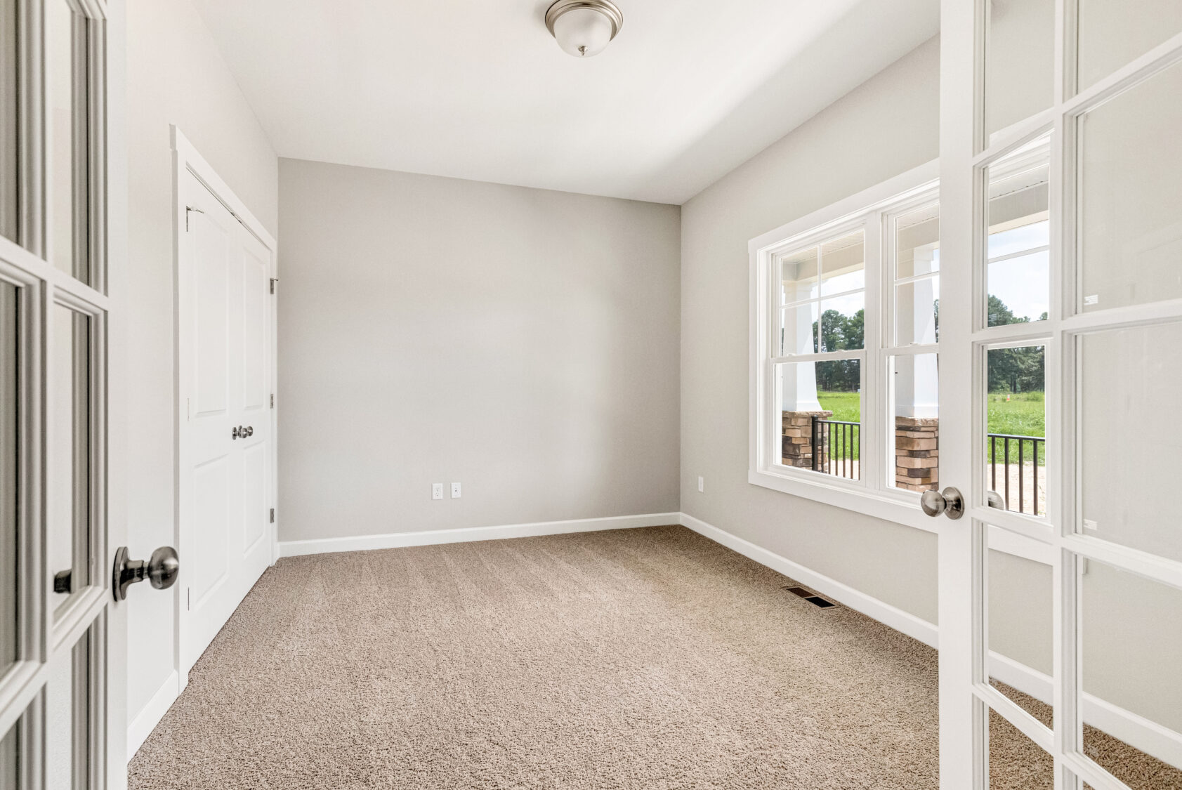 Bright, empty room with beige carpet, white walls, double doors, and a large window showing a green outdoor scene.