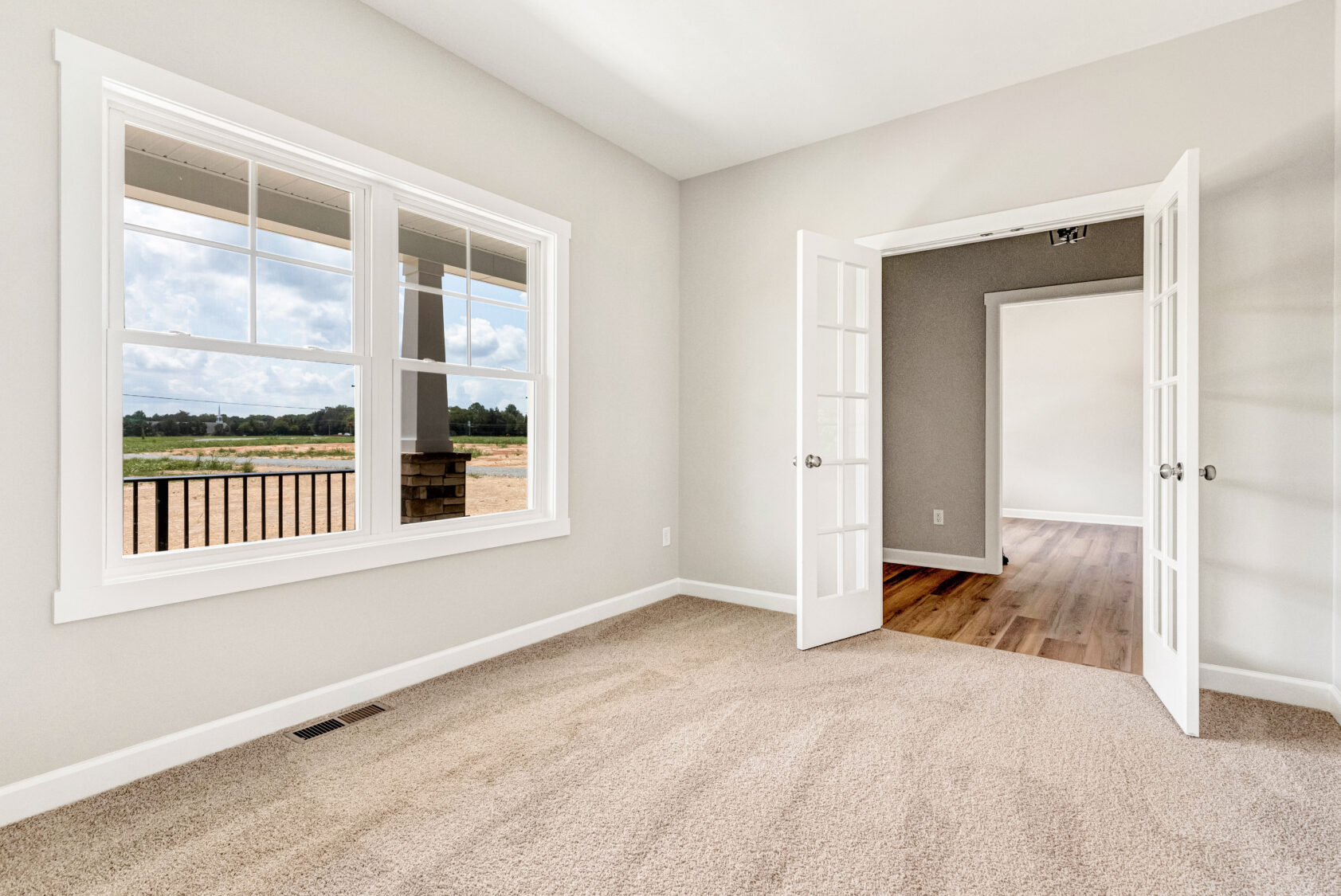 Bright empty room with beige carpet, large window, and open French doors leading to a space with wood flooring.