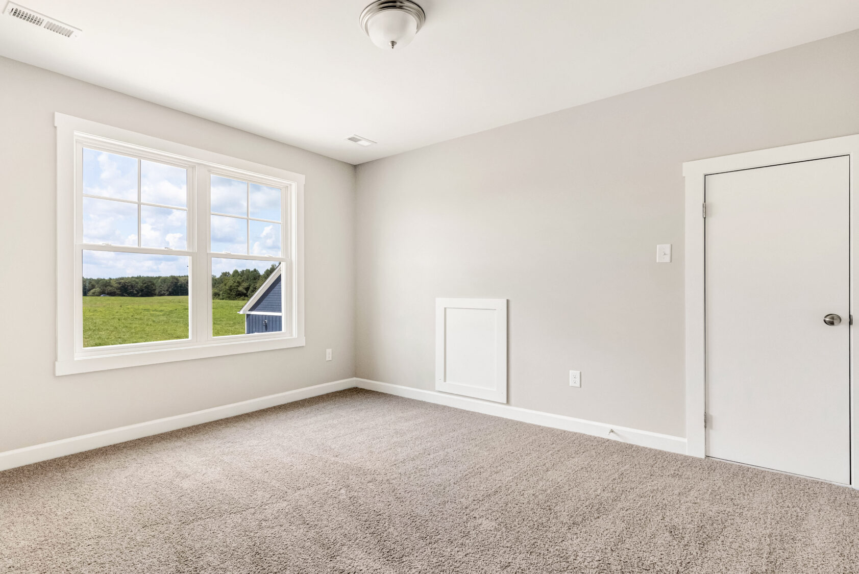Empty room with light gray walls, a window showing a green field, and beige carpeted flooring.
