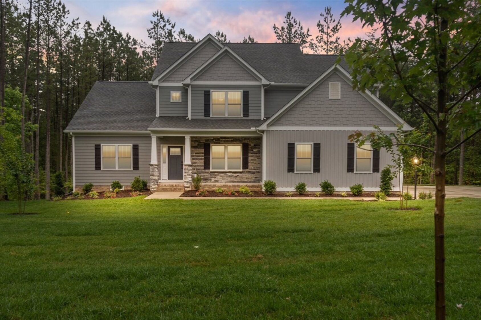 Two-story gray house at Shirley Commons with front lights on, large lawn, shrubs, and trees at sunset.