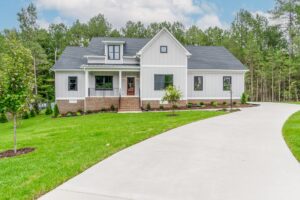 Modern white farmhouse at Shirley Commons with front porch, large windows, and a curved driveway amid green lawn and trees.