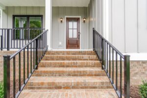 Brick steps with black railings lead to a brown front door at Shirley Commons, a modern gray house with vertical siding.