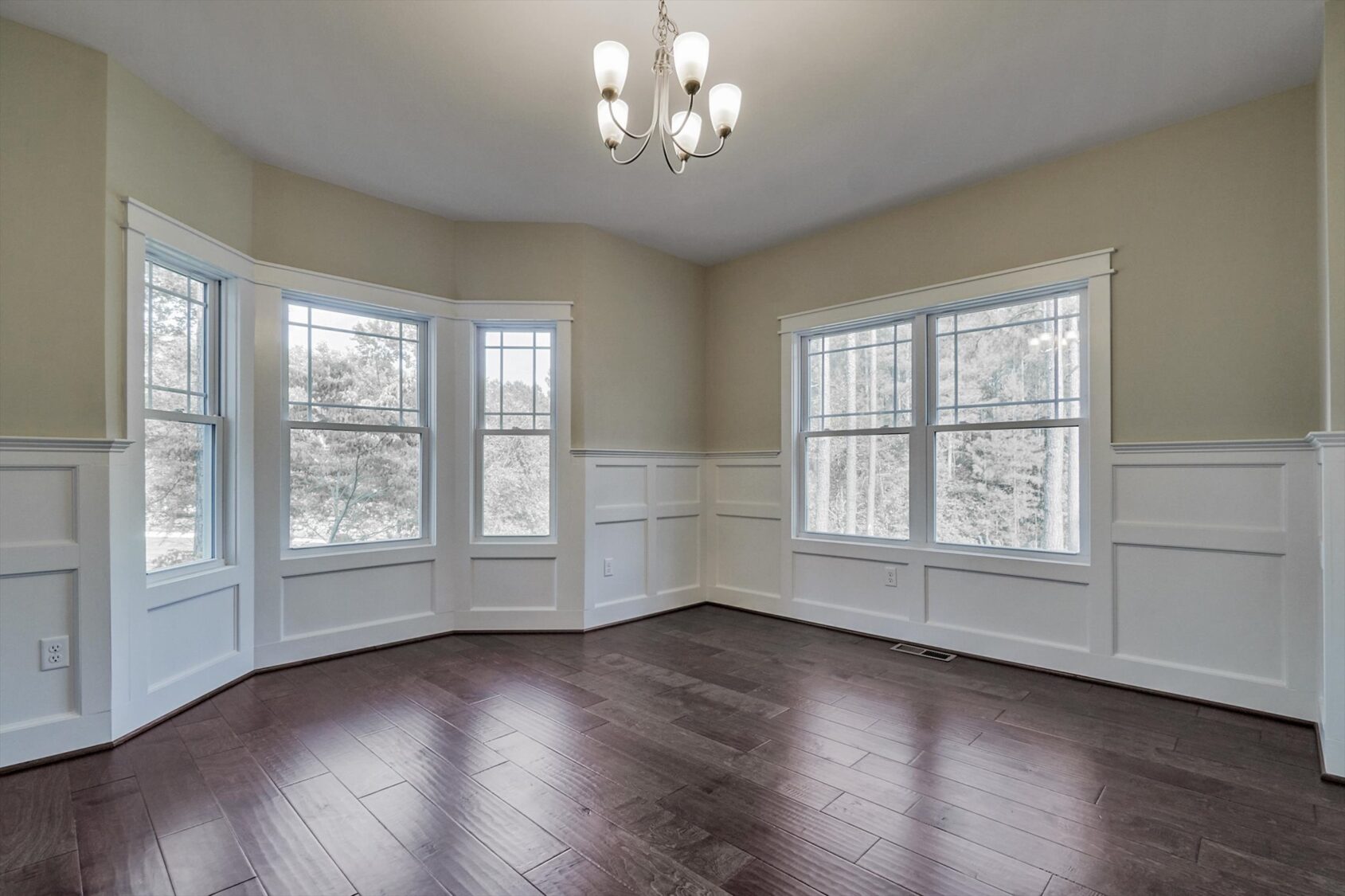 Bright, empty room in Poplar Village with dark wood floors, beige walls, large windows, and modern chandelier.