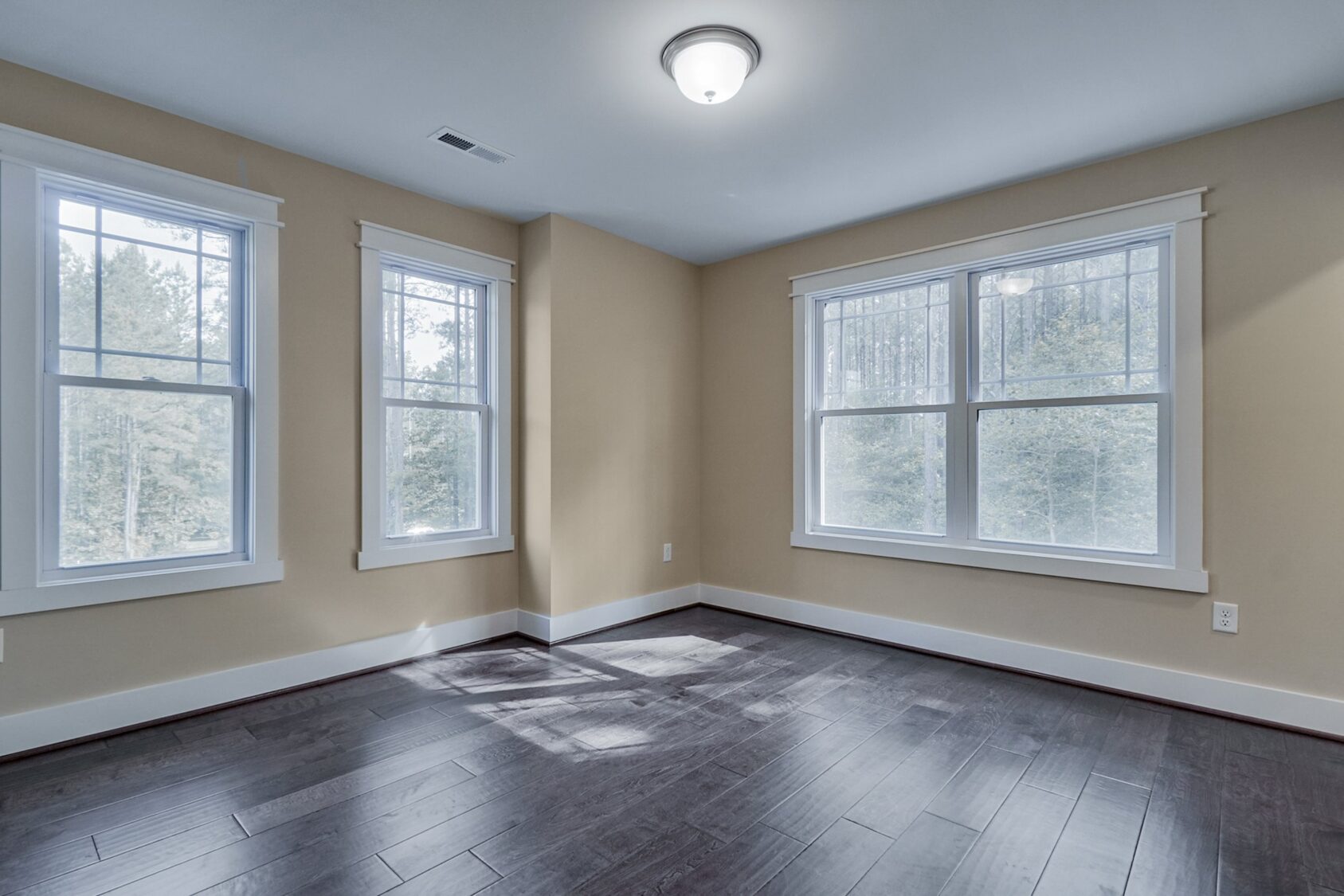 Empty room in Poplar Village with beige walls, dark wood floor, and three large windows letting in natural light.