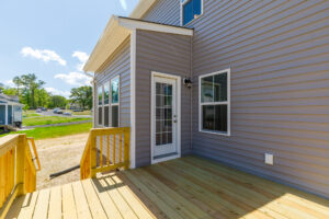 A new wooden deck and steps lead to a white door on a gray home at Lot 28, Poplar Village, on a sunny day.