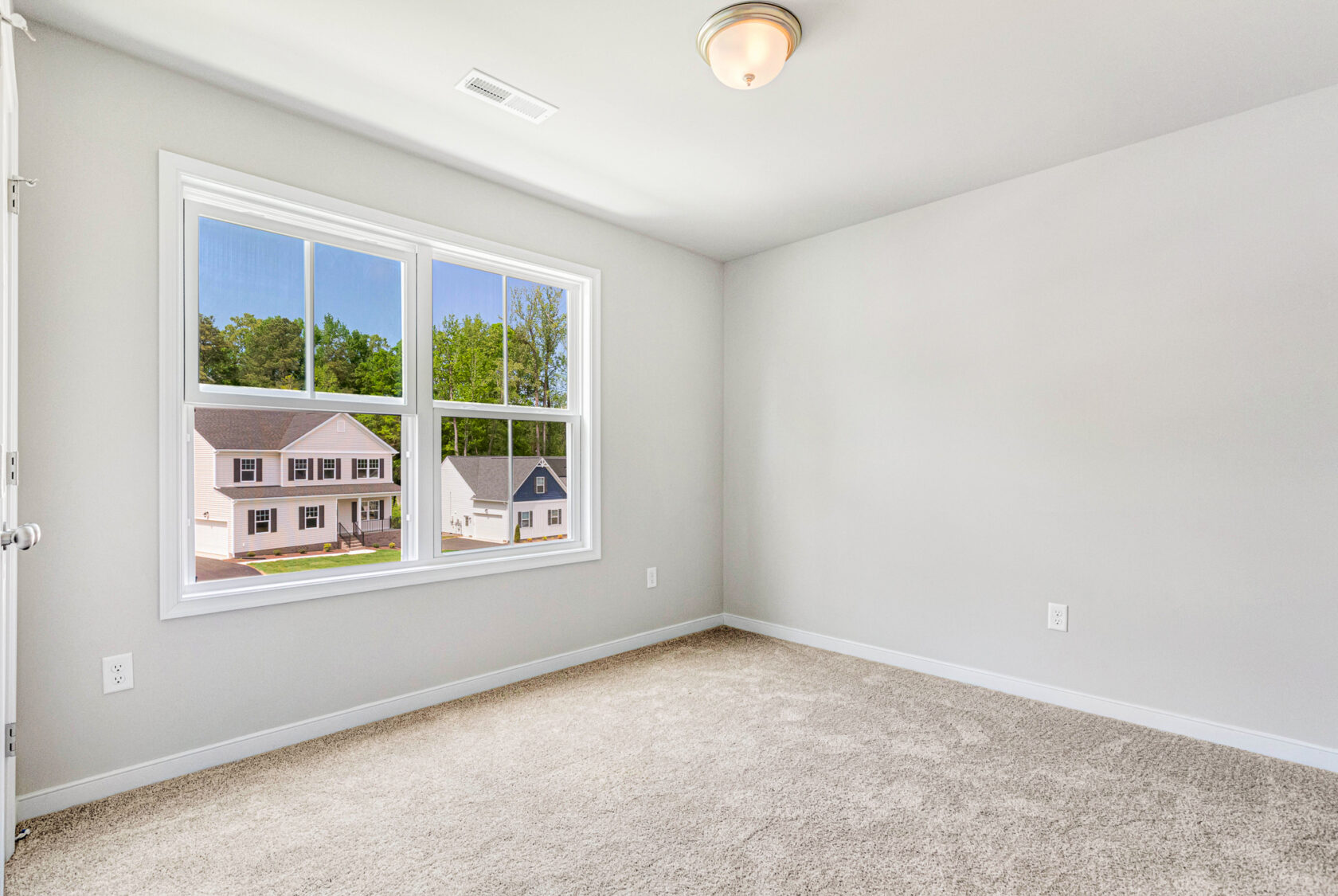 Bright, empty carpeted room in Poplar Village with a large window overlooking houses and trees on a sunny day.