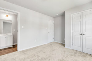 A bright, empty bedroom in Poplar Village with beige carpet, double closets, and a view into a bathroom.