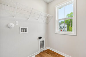 Empty laundry room in Poplar Village Lot 28 with white walls, wire shelf, hookups, window, pet door, and wood floor.