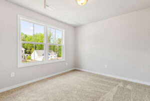 Empty room with beige carpet and gray walls in Poplar Village, featuring a large window with views of Lot 28 and greenery.