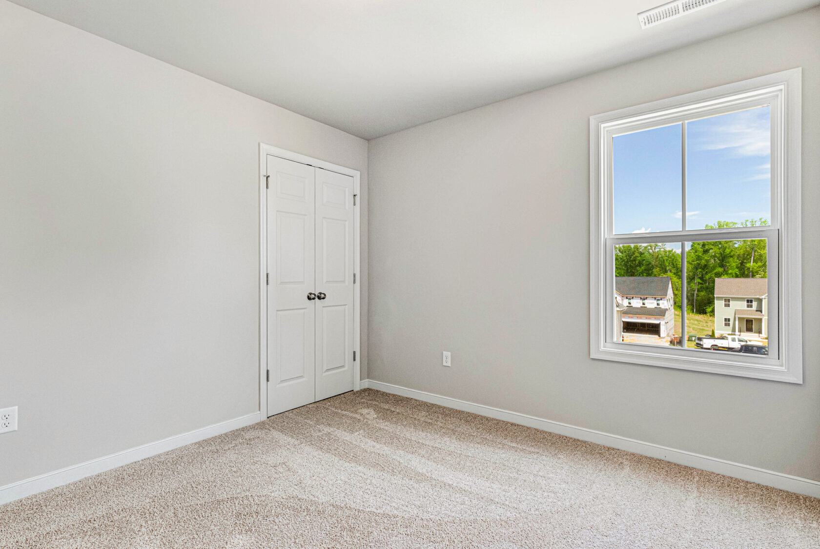 Empty room in Poplar Village with beige carpet, white walls, double door closet, and window overlooking Lot 28.