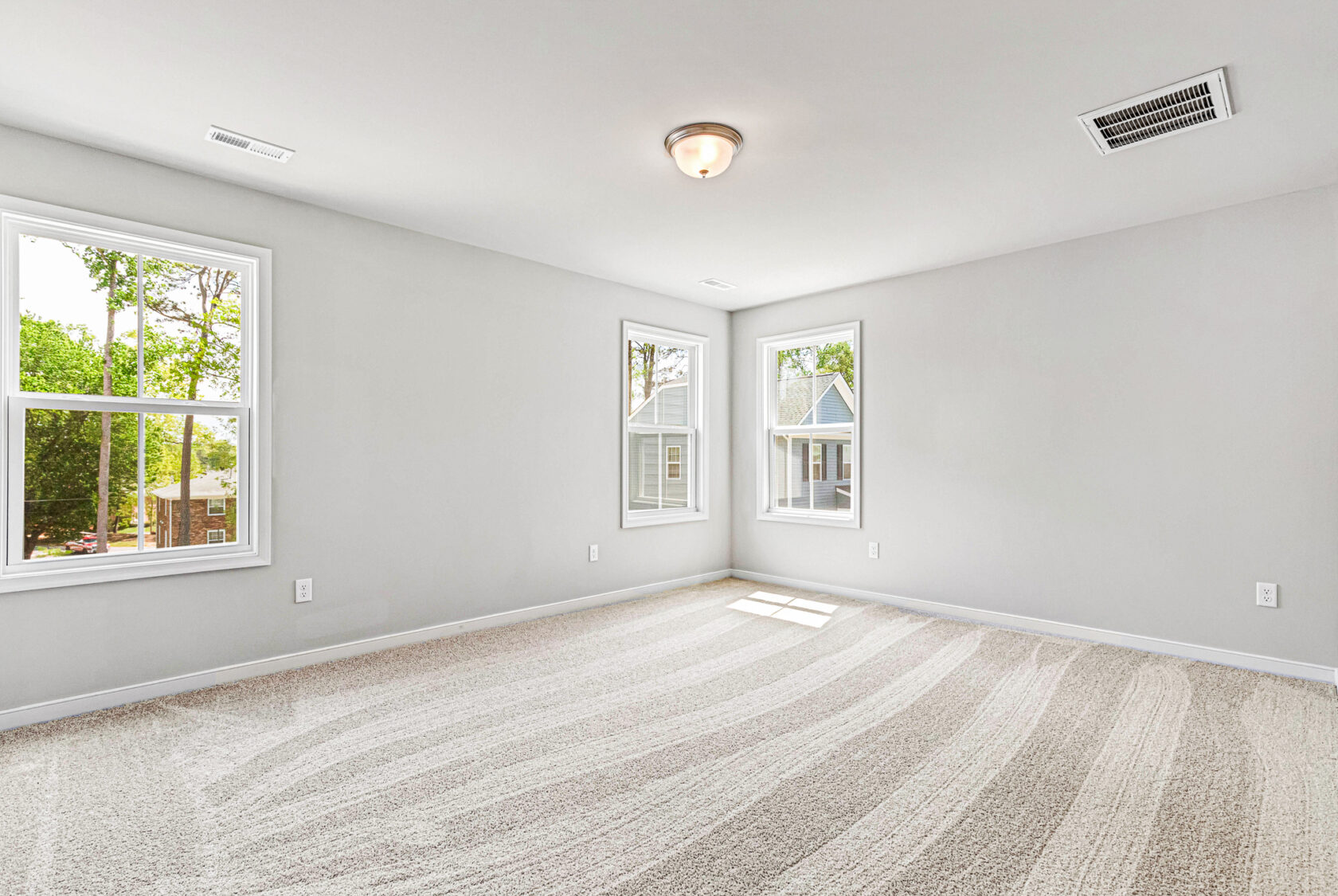 Bright, empty carpeted room in Poplar Village with white walls, ceiling light, and windows overlooking houses and greenery.