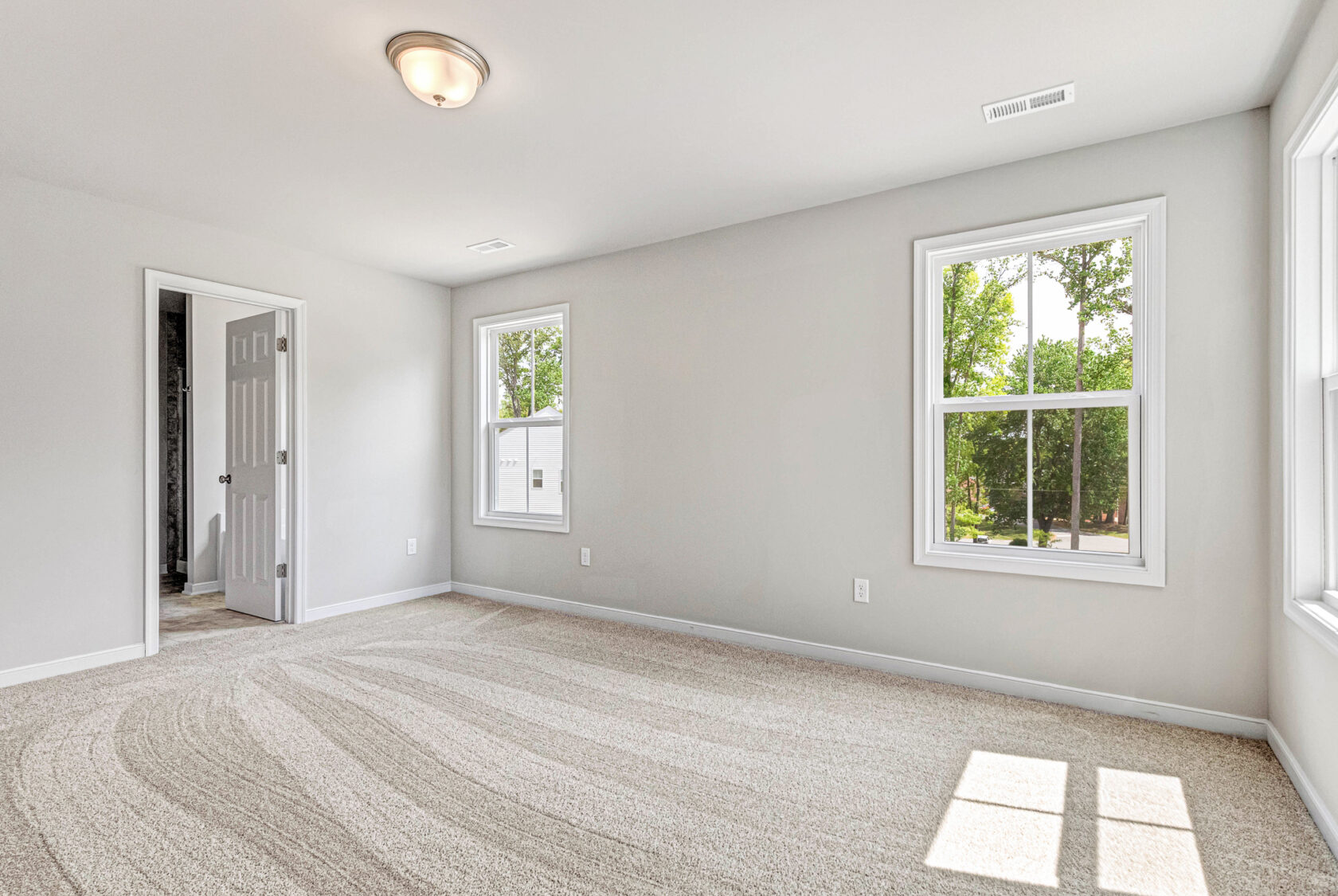 Bright, empty bedroom in Poplar Village with beige carpet, white walls, two windows, and open door to bathroom.
