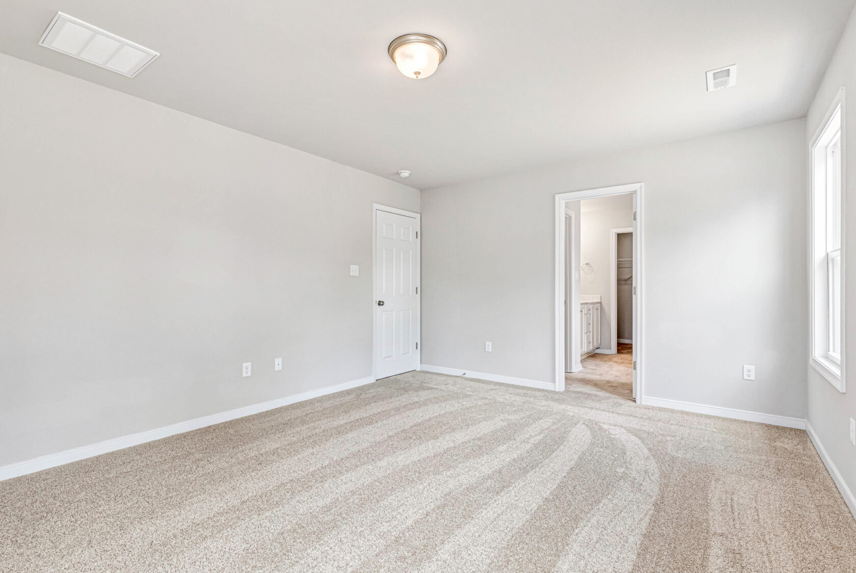 Empty, bright carpeted room in Poplar Village with white walls, ceiling light, and doors to hallway and bathroom—Lot 28.