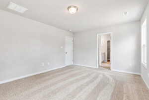 Empty, bright carpeted room in Poplar Village with white walls, ceiling light, and doors to hallway and bathroom—Lot 28.