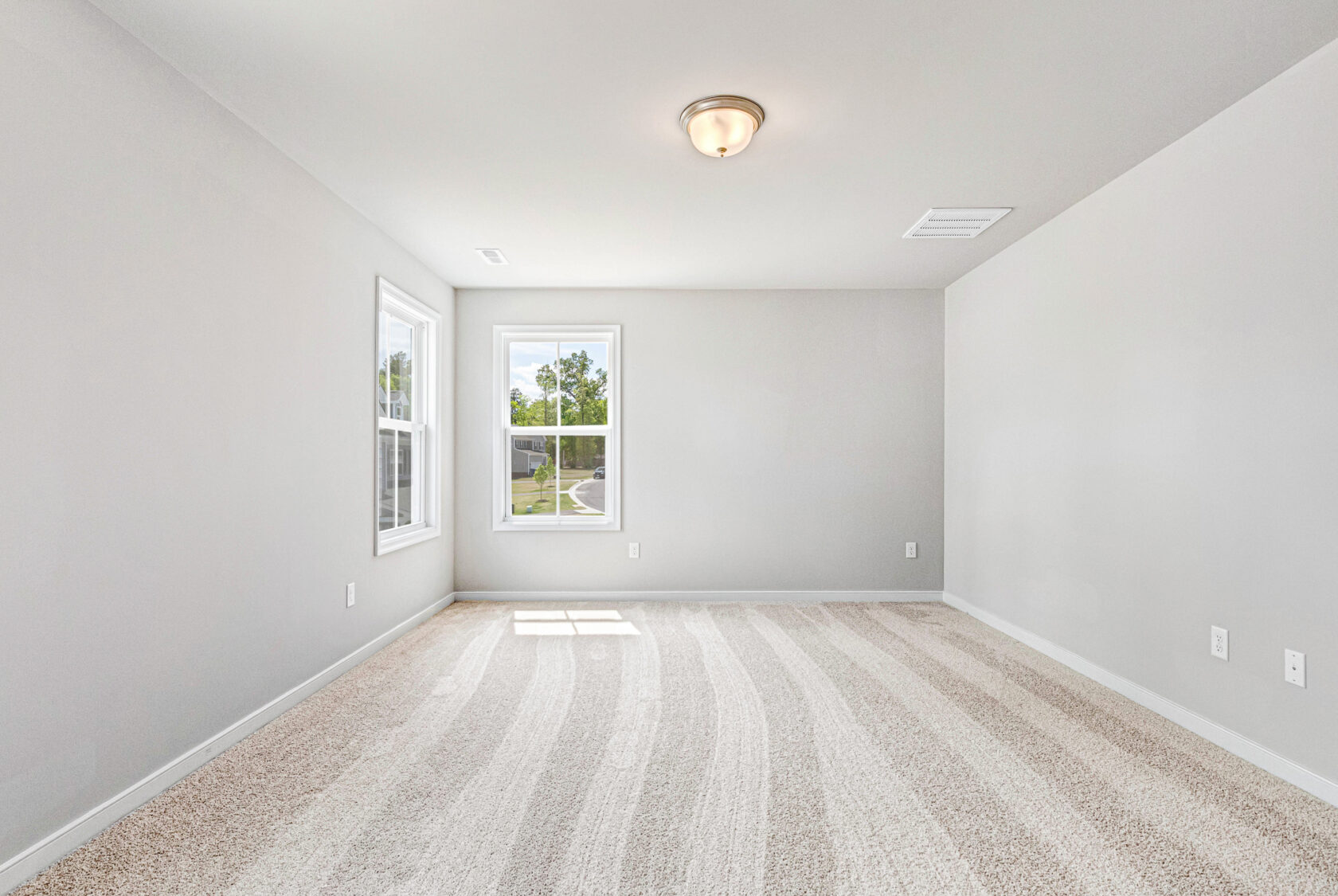 Bright, empty room in Poplar Village, Lot 28 features beige carpet, light gray walls, two windows, and a ceiling light.