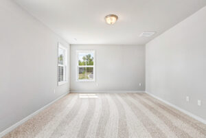 Bright, empty room in Poplar Village, Lot 28 features beige carpet, light gray walls, two windows, and a ceiling light.