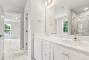Modern bathroom with double sink vanity, large mirror, and view into Lot 28 of Poplar Village’s bright carpeted bedroom.