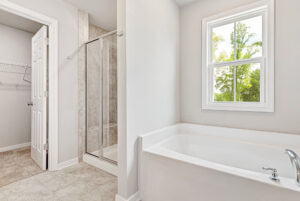 Bright bathroom in Lot 28, Poplar Village with a large tub, glass shower, closet door, and window with tree views.