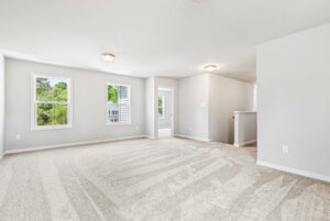 Spacious, empty carpeted room in Poplar Village with white walls, large windows, and a door to another area.