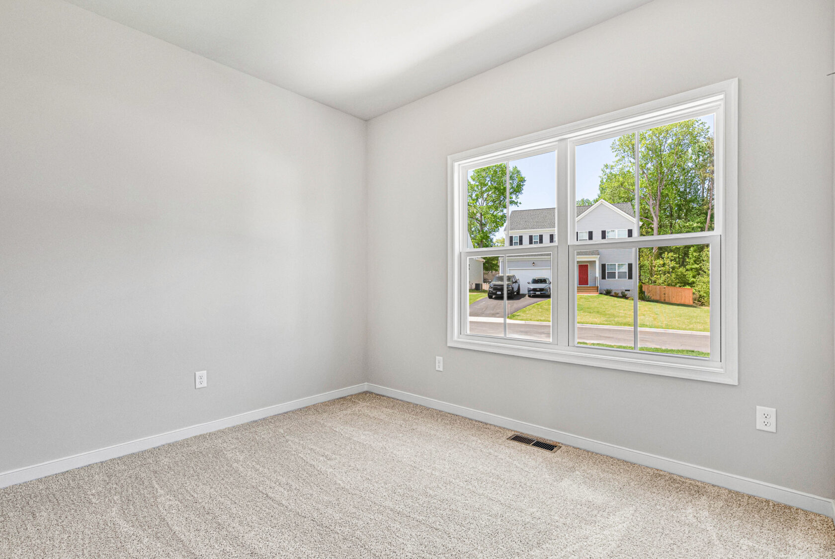 Empty, carpeted room in Poplar Village with light gray walls and a large window overlooking Lot 28 and leafy trees.