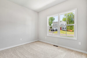 Empty, carpeted room in Poplar Village with light gray walls and a large window overlooking Lot 28 and leafy trees.