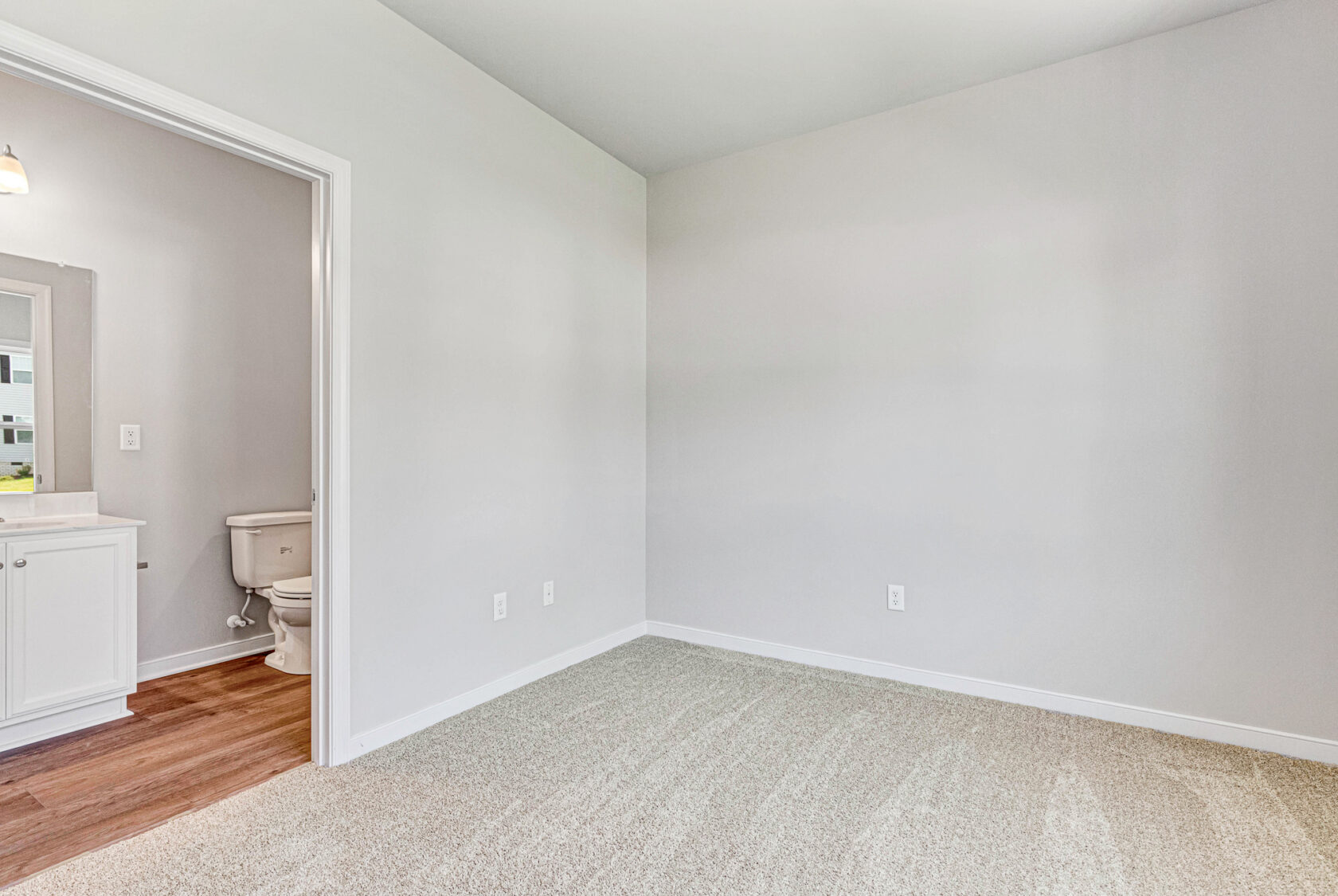 Empty beige-carpeted room with white walls in Poplar Village, next to a bathroom with toilet and sink visible. Lot 28.