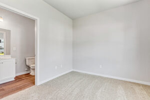 Empty beige-carpeted room with white walls in Poplar Village, next to a bathroom with toilet and sink visible. Lot 28.
