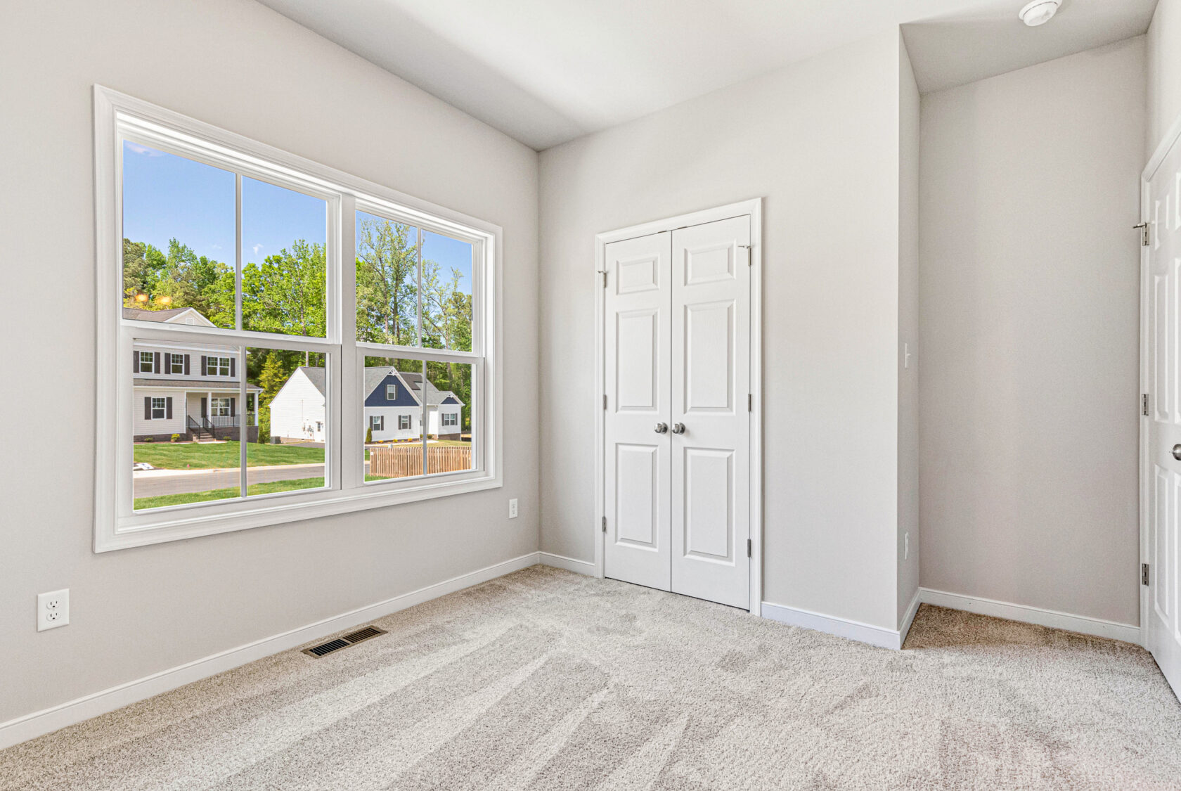 Bright, empty room with beige carpet, large window, and double-door closet in Poplar Village’s Lot 28; view of houses outside.