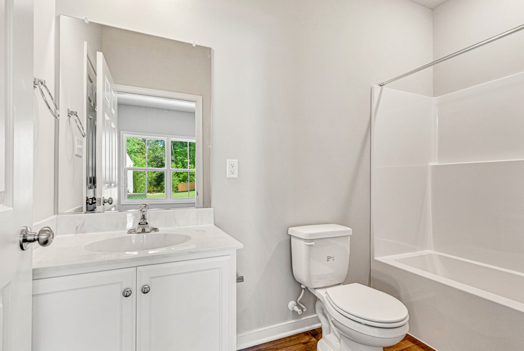 White bathroom with a sink, mirror, toilet, bathtub, and a window overlooking Poplar Village greenery. Lot 28.