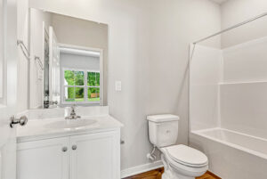 White bathroom with a sink, mirror, toilet, bathtub, and a window overlooking Poplar Village greenery. Lot 28.