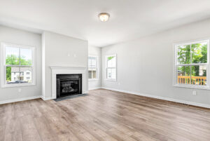 Bright, empty living room with wood floors in Poplar Village, Lot 28; fireplace and large windows let in natural light.