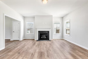 Bright, empty living room in Poplar Village Lot 28 with light wood floors, a black fireplace, and three sunny windows.