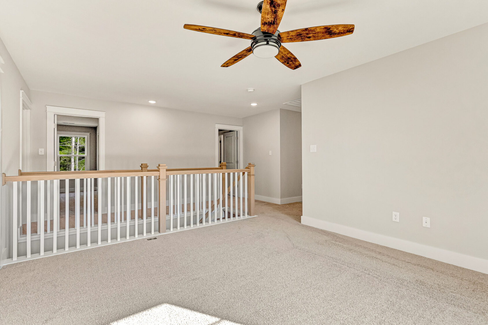 Bright carpeted room with tan walls in Somerset Hill, wooden ceiling fan, and white railing overlooking hallway.
