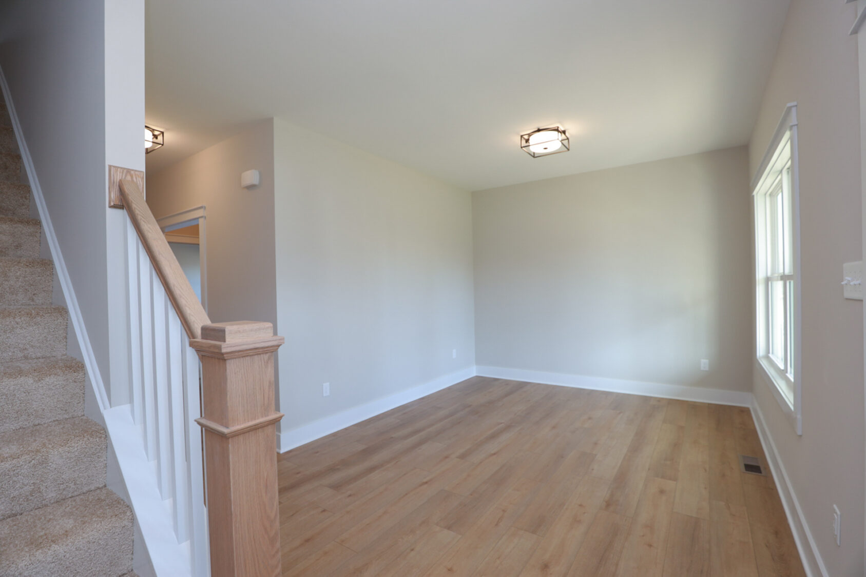 Empty living room with wood floors in Somerset Hill, beige walls, a window, and carpeted stairs with white railing on left.